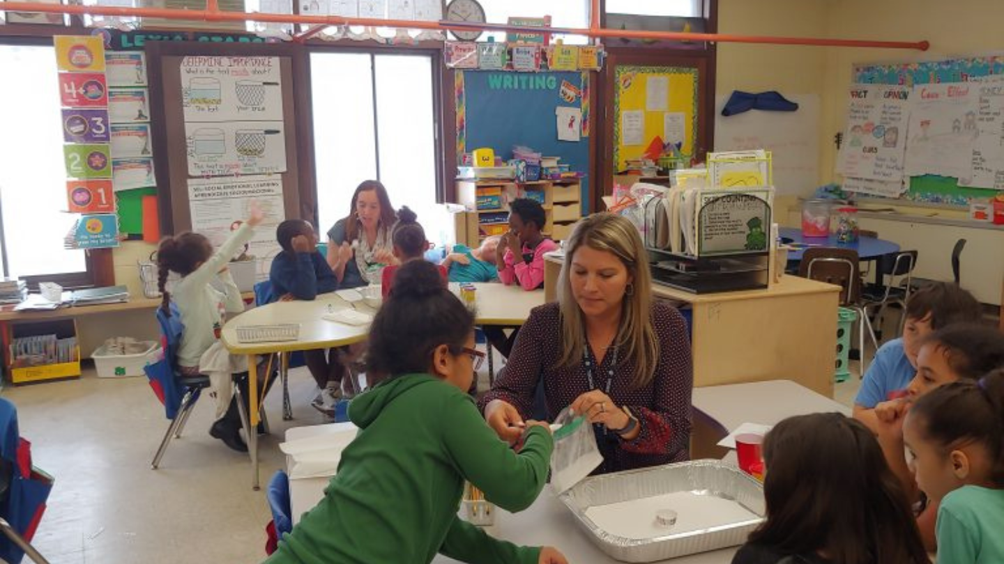 Two teachers sit at desks facilitating a program with elementary students. 