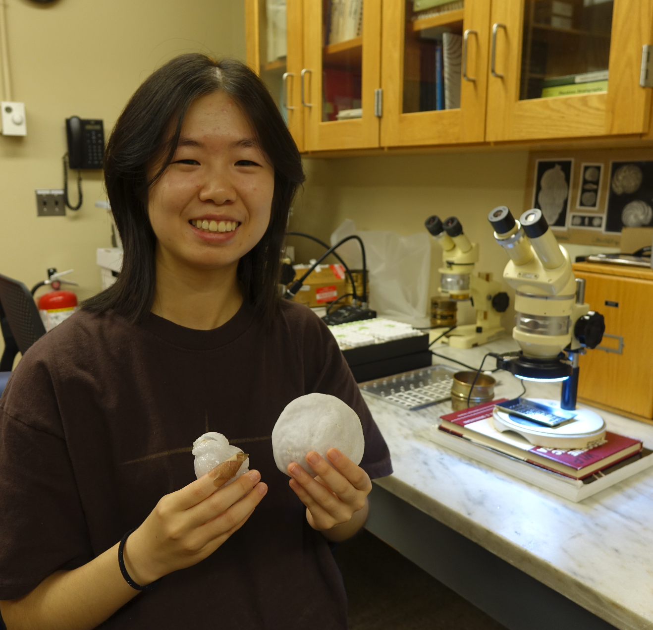 Claire in the lab, smiling beside the microscope while holding 3D print models of microfossils. 