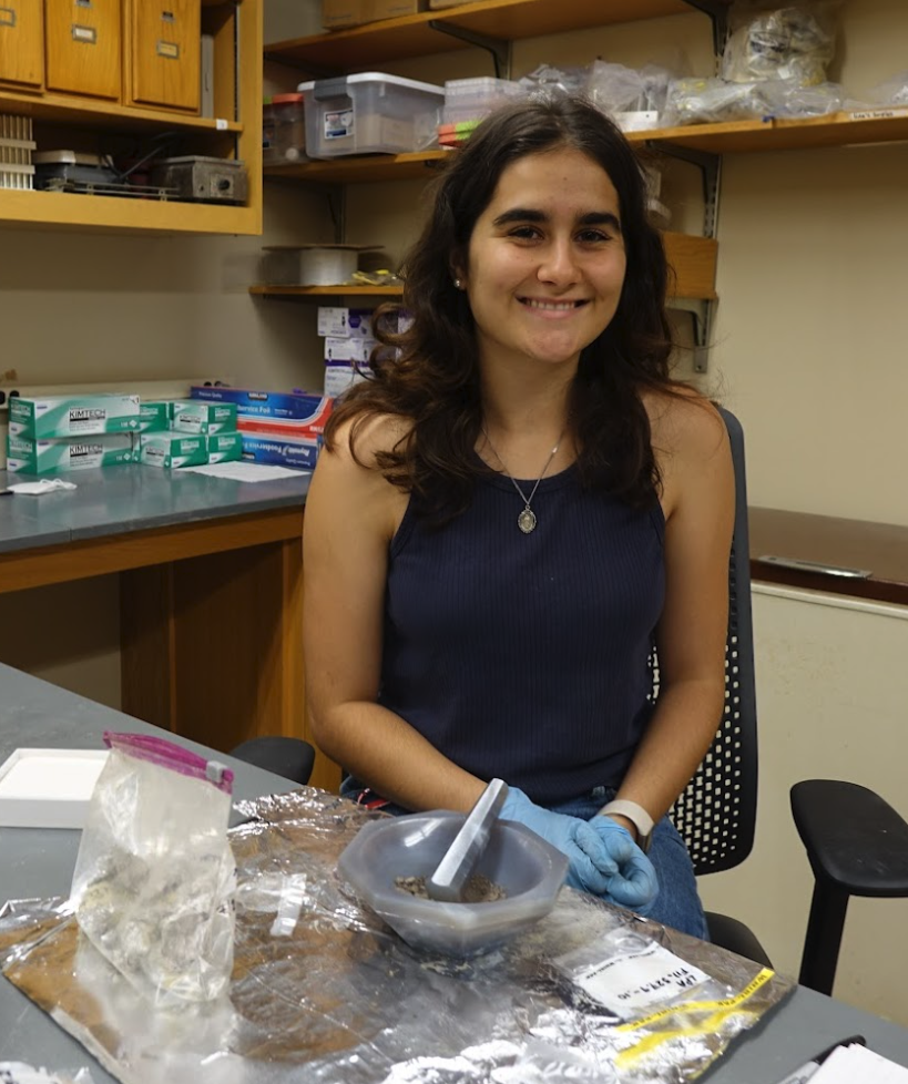 Isabel smiling in the lab beside a mortar and pestle with samples. 