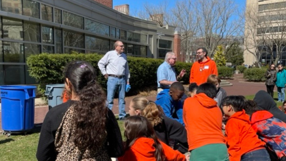 A group of third and fourth grade students sit outside facing the DEEPS faculty for the volcano demonstration.