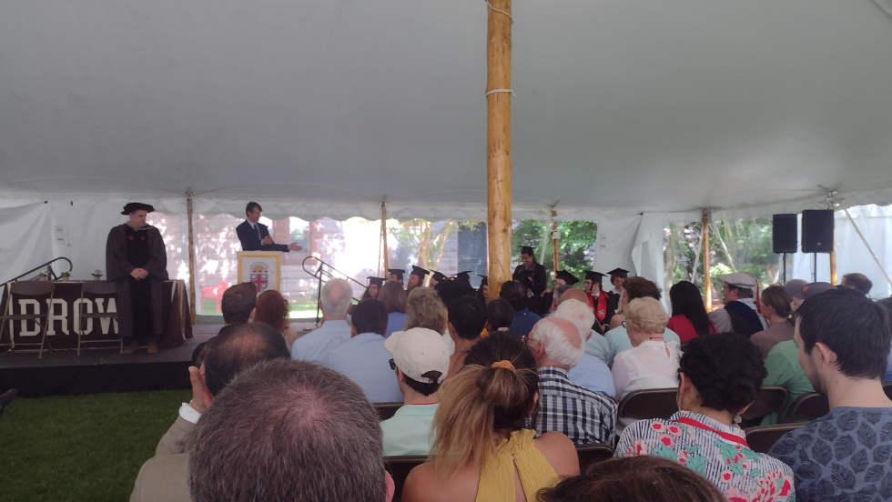 A wide photo of the commencement event, with families watching the Chair speak to the graduates.