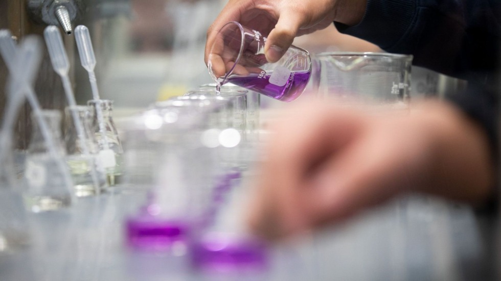 Hands pouring purple liquid from beakers in a chemistry lab.