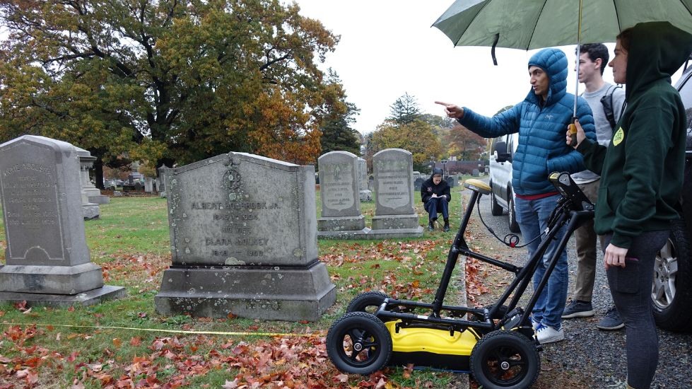 Hannah Krueger stands with students in the cemetery, holding an unbrella over the GPR unit (which looks like a small cart). 