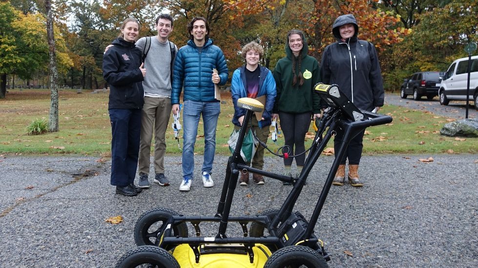 Participants stand to smile for a photo, with the GPR in front of them and the cemetery behind them. 