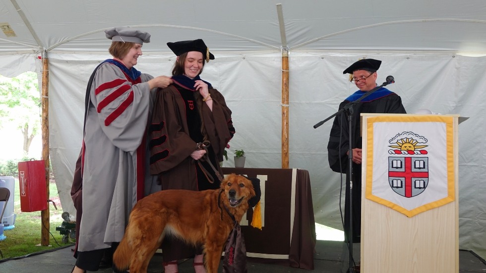 Karen Fischer putting the doctoral hood on a graduate, while her dog joins in
