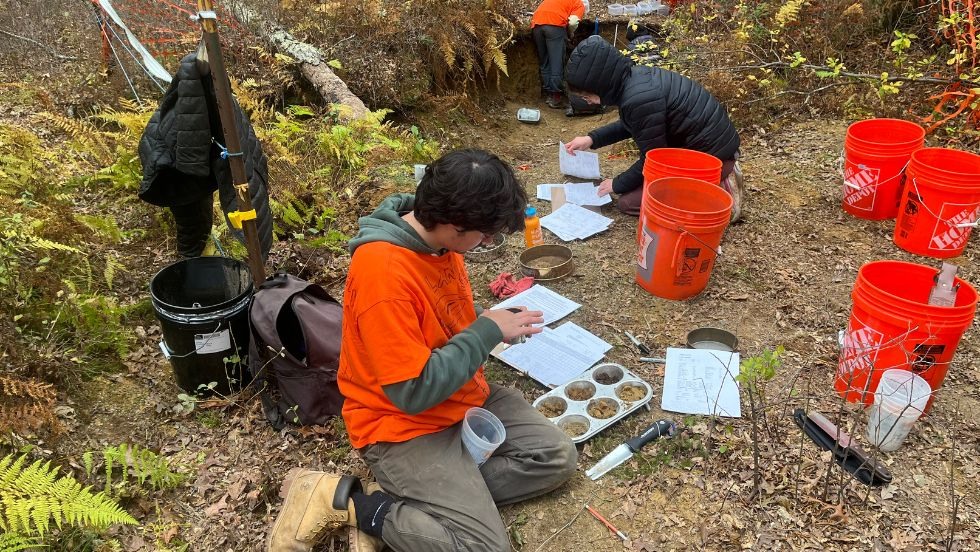 a student examines a soil sample seated beside a hole where fellow students are digging. 