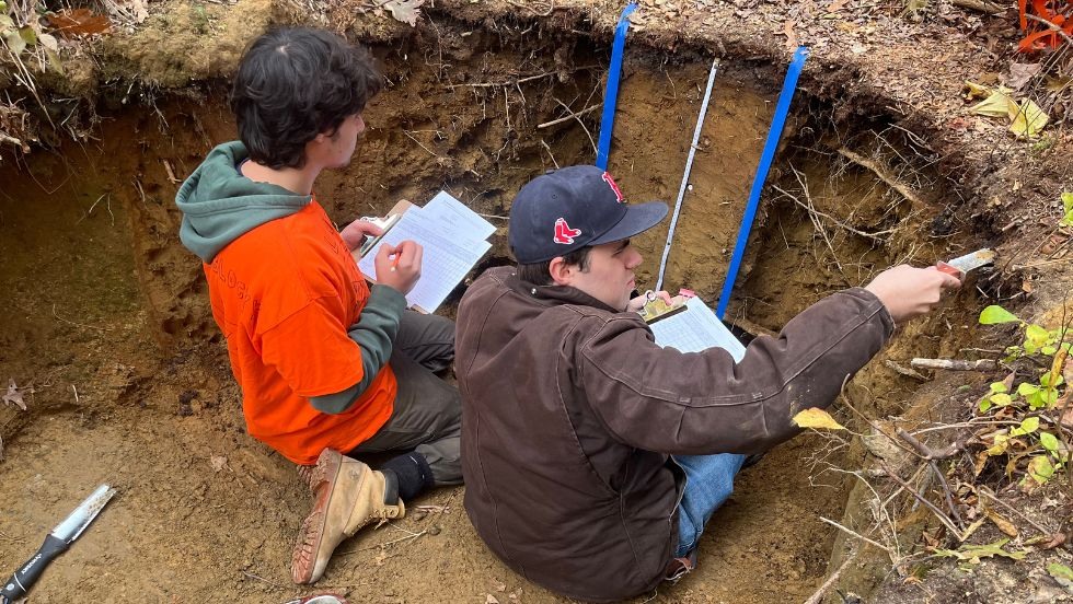 two students seated in a hole taking soil samples