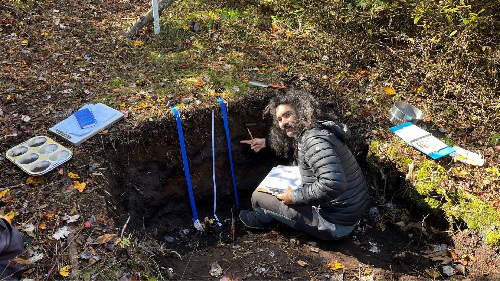 a graduate student smiles while sitting in a hole and pointing at the soil