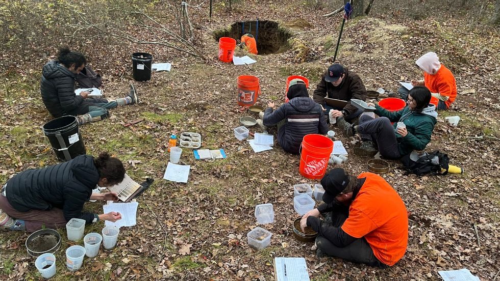 students seated on the ground around a hole measuring soil samples