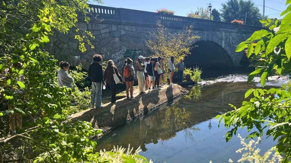 students standing on a riverbank beside a stone bridge