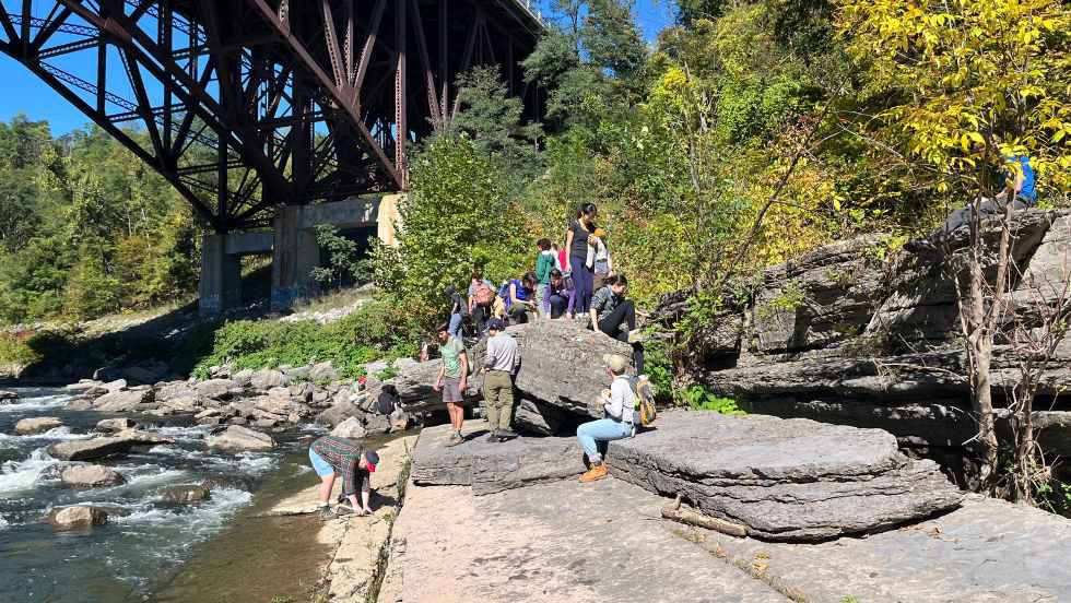 students sitting on rocks beside a creek, under an old steel bridge 