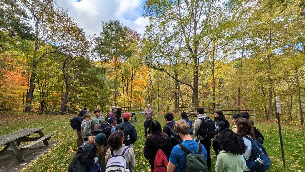 Students circling Proffesor Hodgin as he explains a lesson, outdoors in a forest area