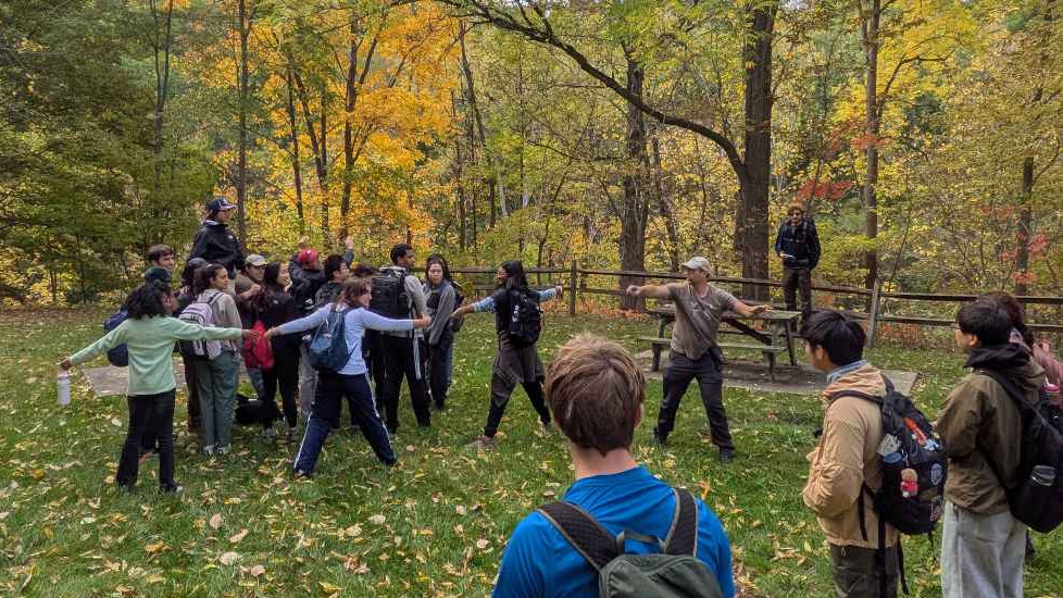 Students forming groups and geologic-style formations while Proffesor Hodgin as he explains a lesson, outdoors in a forest area